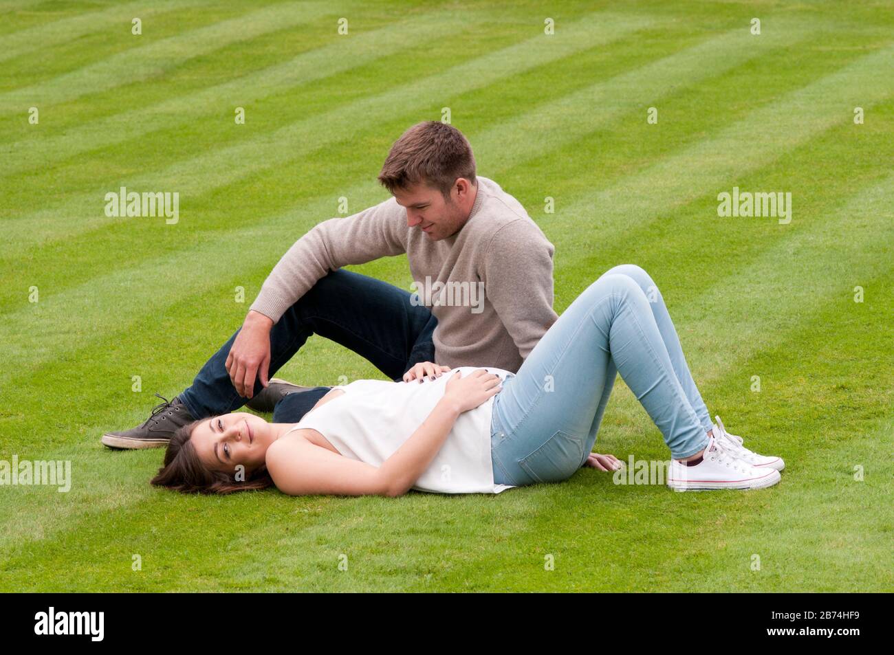 Beautiful pregnant young woman lying down on the grass with her partner sitting next to her Stock Photo