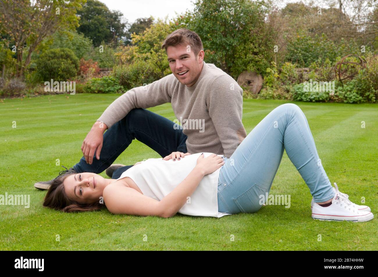 Beautiful pregnant young woman lying down on the grass with her partner sitting next to her Stock Photo