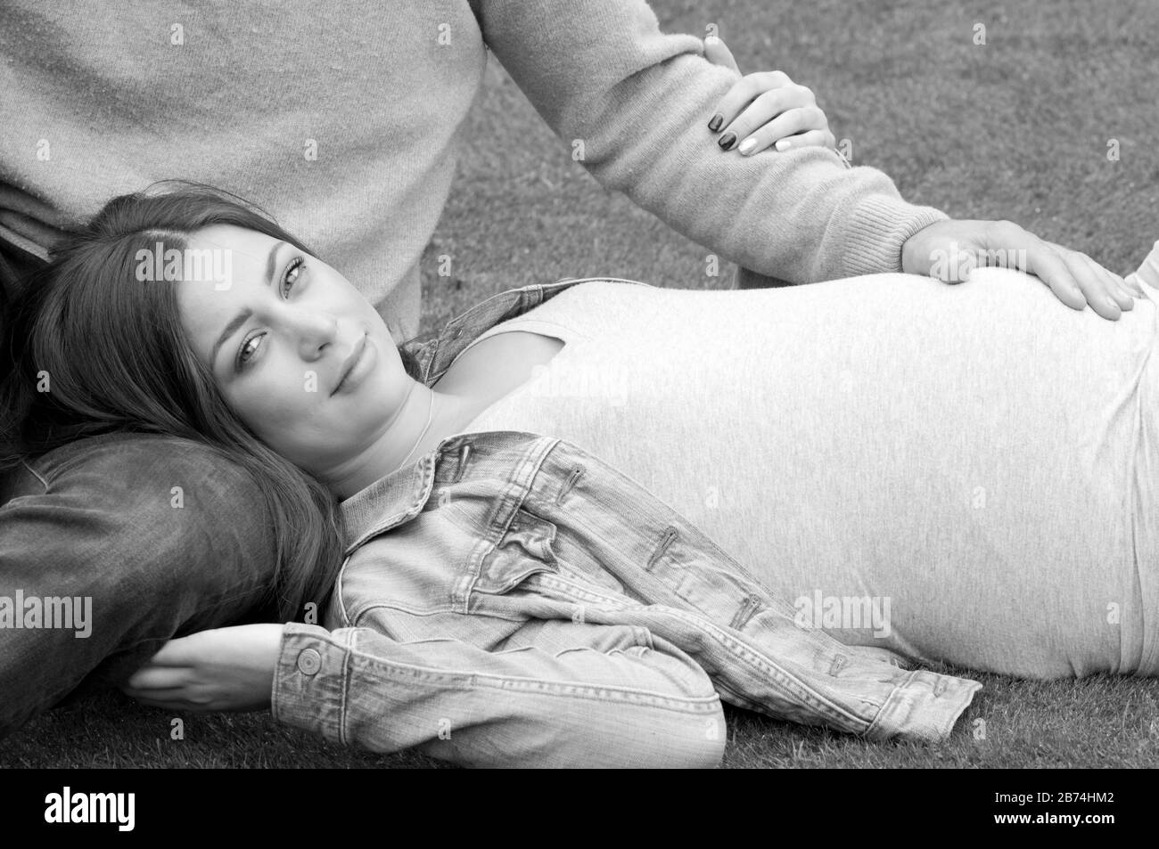 Black and white, close up, beautiful pregnant young woman lying down on the grass with her partner sitting next to her Stock Photo