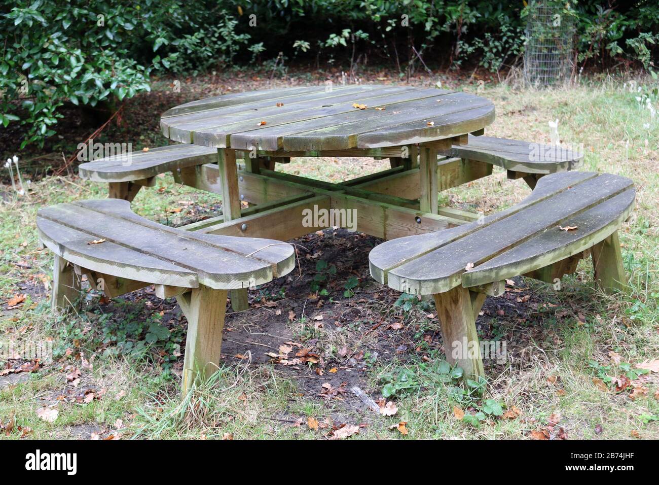 Round wooden picnic table with rounded seats on grass with soil underneath and shrubs in the background. Stock Photo