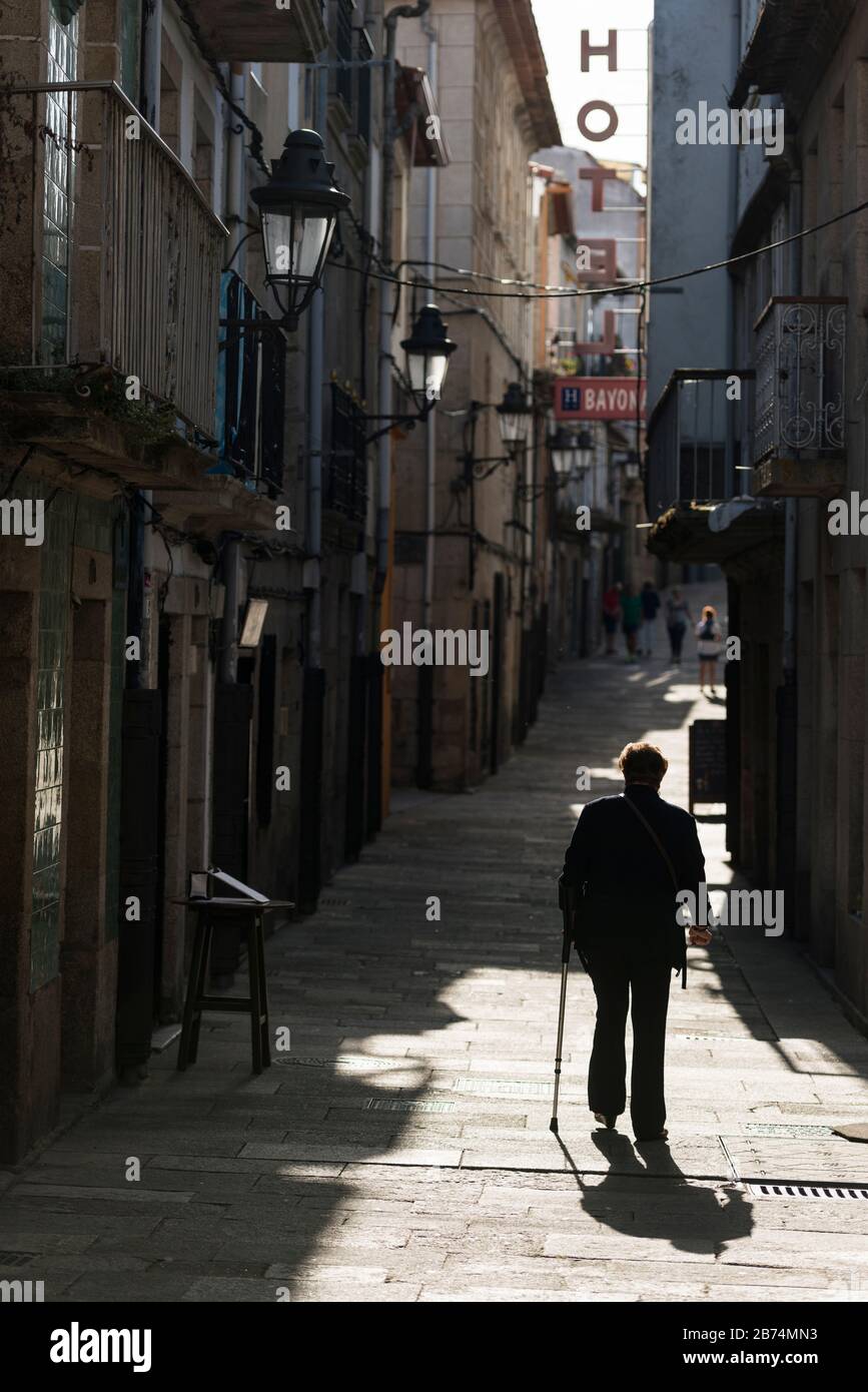 An elderly woman using a walking stick walks up a paved alleyway towards a hotel in the old town area of Baiona, Spain Stock Photo