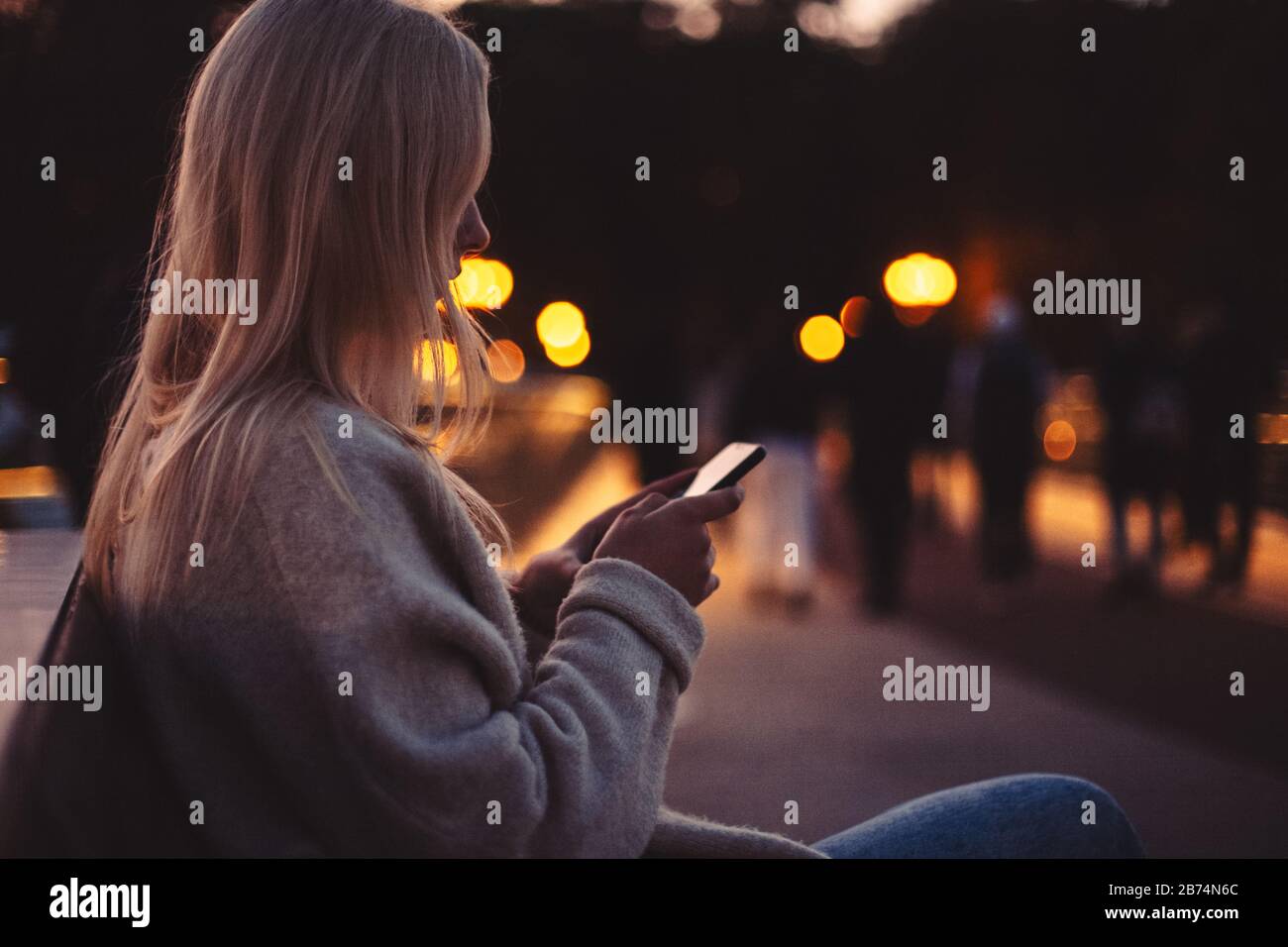 Young woman using smart phone while sitting on bench in city at dusk Stock Photo
