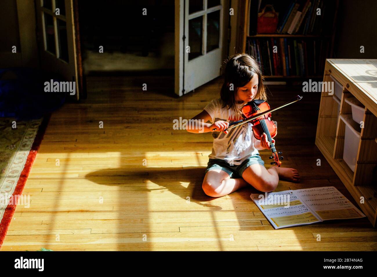 A small child sits alone in a patch of sunlit playing violin Stock Photo