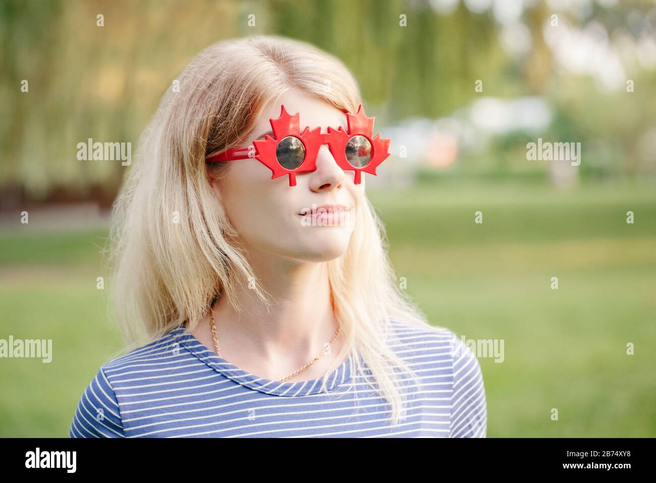 Beautiful blonde Caucasian young girl wearing canadian maple leaf sunglasses. Happy woman with funny red glasses. Citizen female celebrating Canada Da Stock Photo