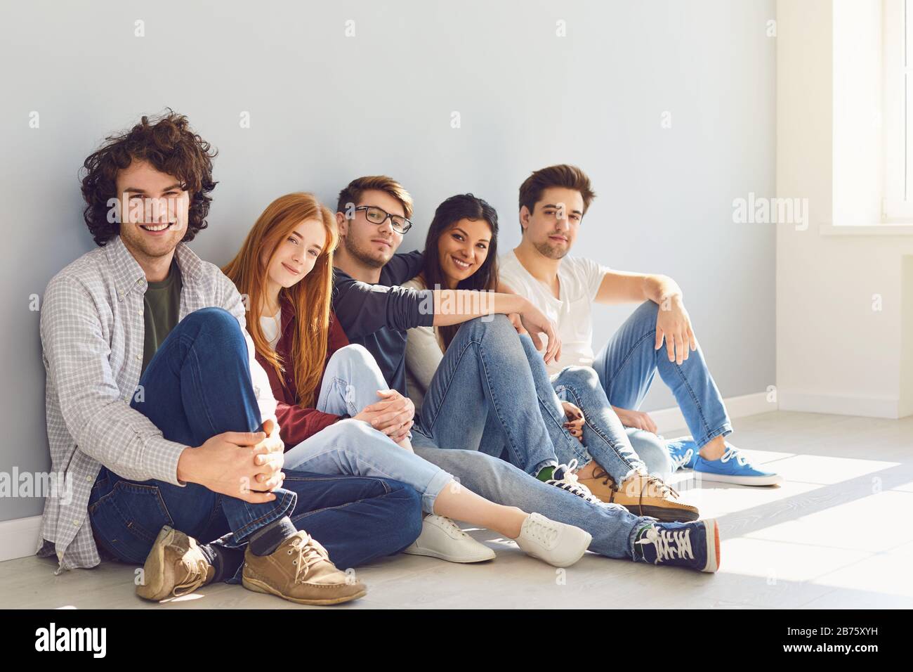 A group of friends is sitting on the floor in a room on a gray background. Stock Photo