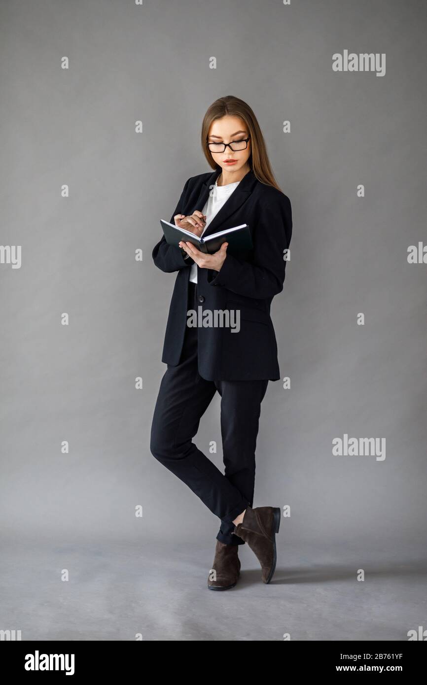 Young pretty girl in business style clothes stands on a gray background with a notebook and a pen in her hands Stock Photo