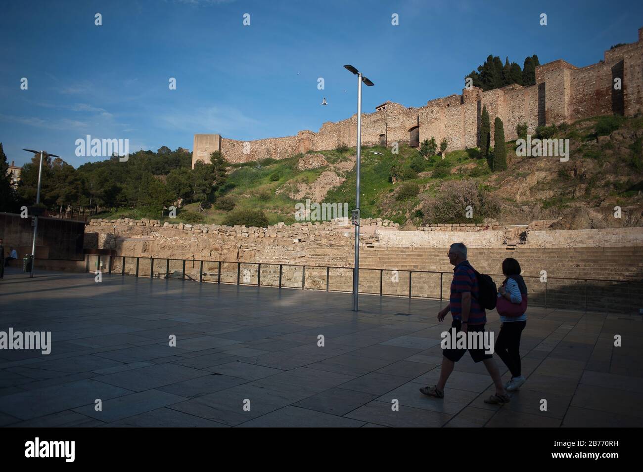 Malaga, Spain. 13th Mar, 2020. People walk on the street as the facade of Roman theatre is being closed to visitors.Spanish Prime Minister Pedro Sánchez has declared the emergency state in Spain because of Covid-19 outbreak, after the raise of number of cases of people infected with Covid-19 in the country. Spanish authorities have closed museums and monuments to control the expansion of Covid-19 outbreak. The city of Malaga is the first in Andalusia with the most number of people infected. Credit: SOPA Images Limited/Alamy Live News Stock Photo