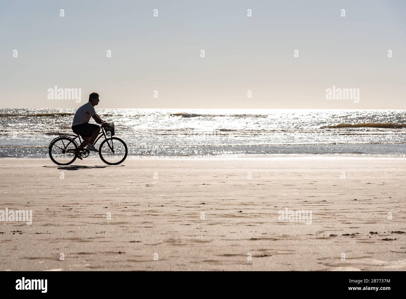 Silhouette of man riding bicycle on a beach with the sun reflecting in the sea and a clear sky Stock Photo