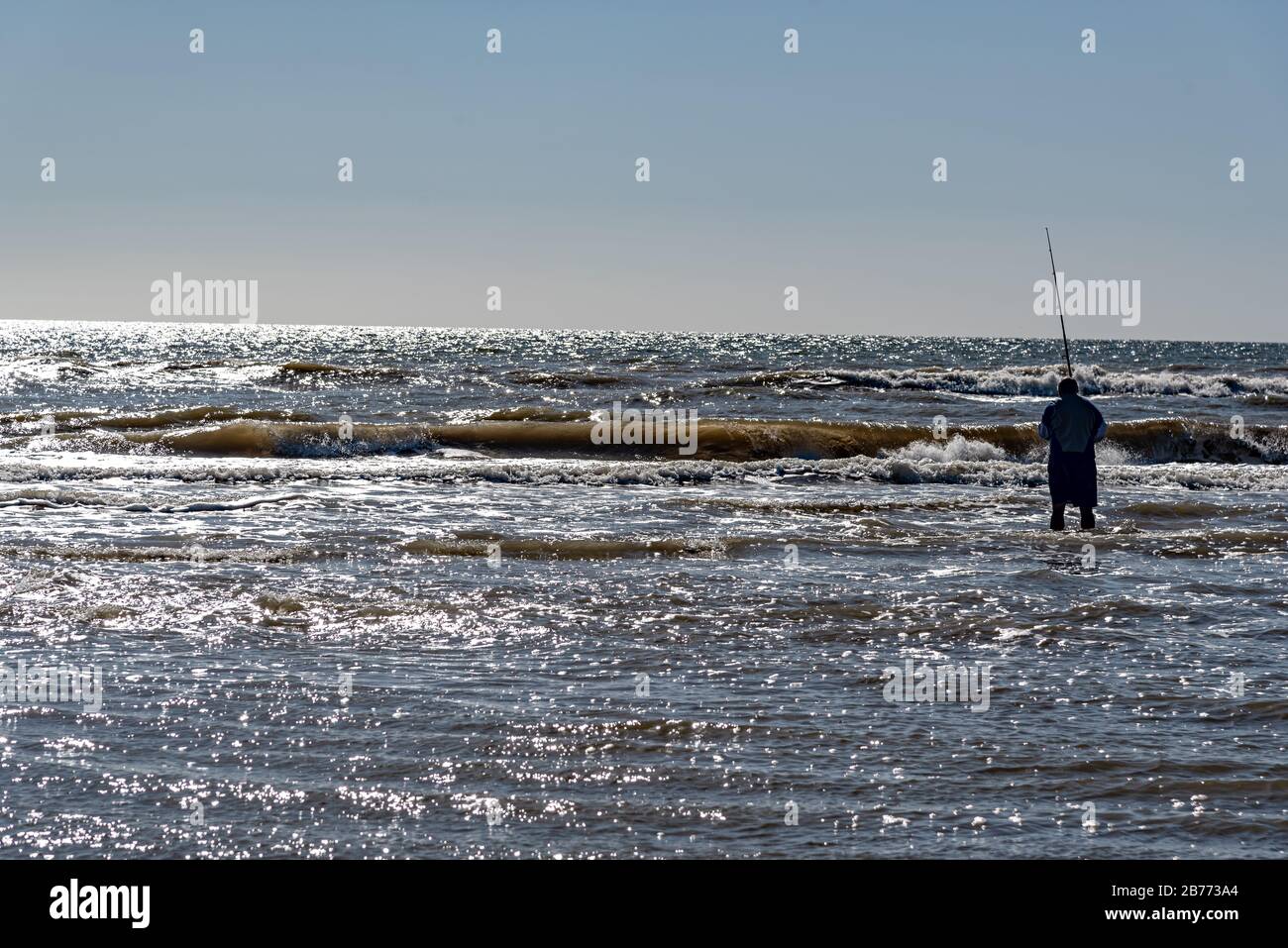 Silhouette of a man fishing alone on the beach in the water with a view of the horizon Stock Photo