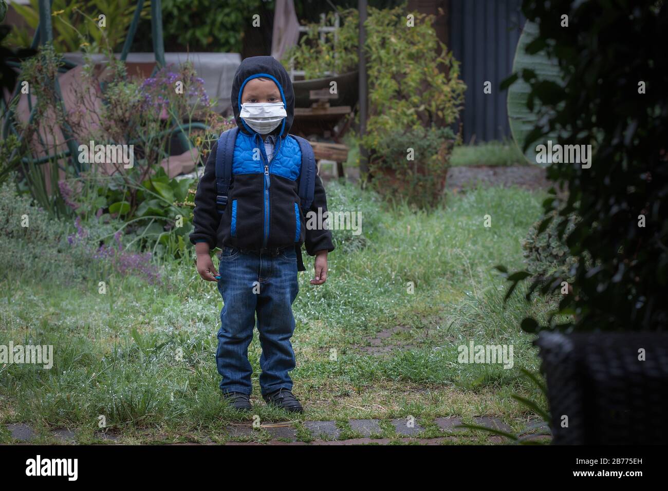 A young boy wearing a backpack, face mask, hoodie jacket, and rain boots, standing outdoors while its raining. Stock Photo