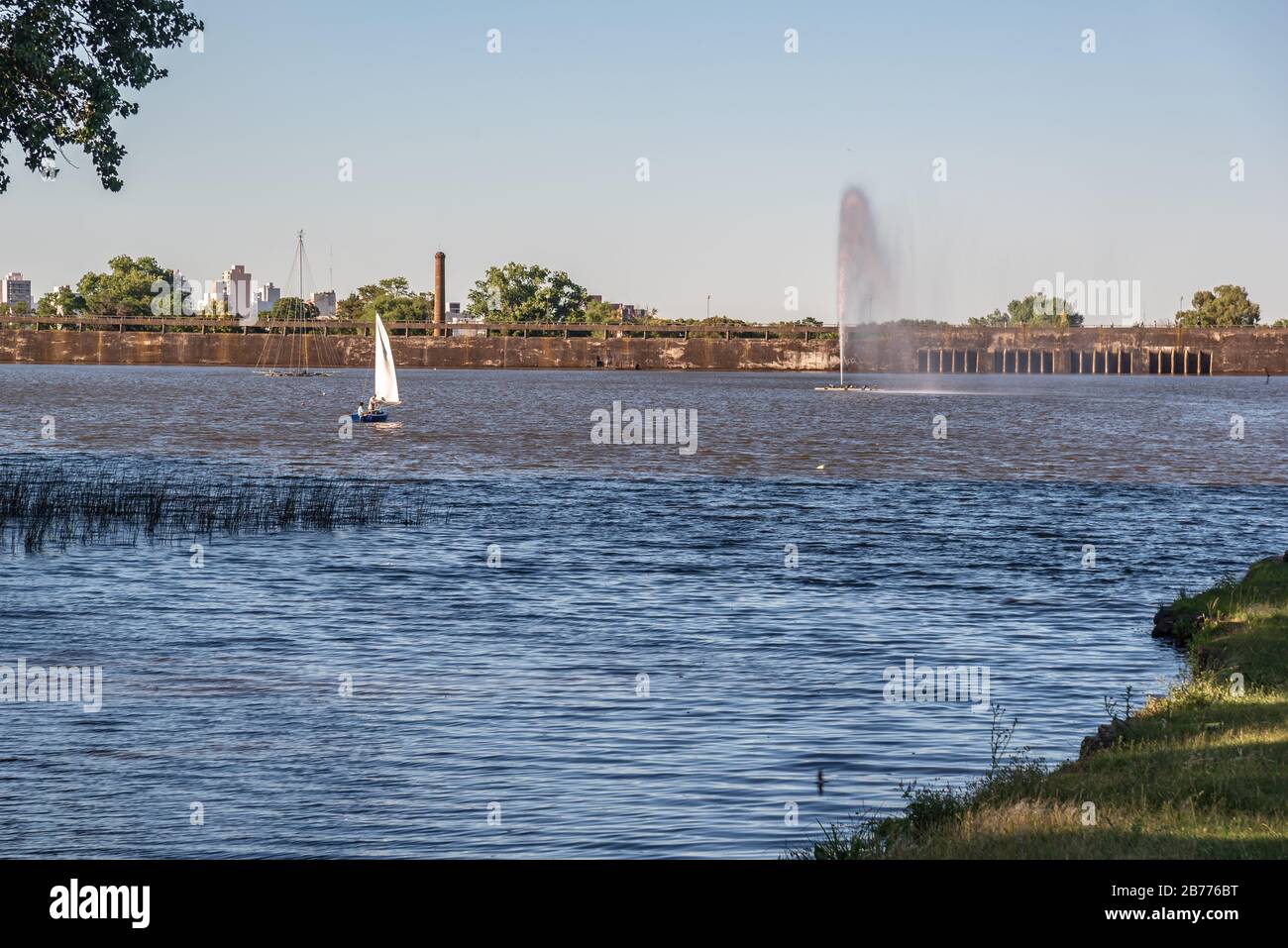 View of a blue lake with a city in the back a sailboat and a spring of water high up from the shore Stock Photo