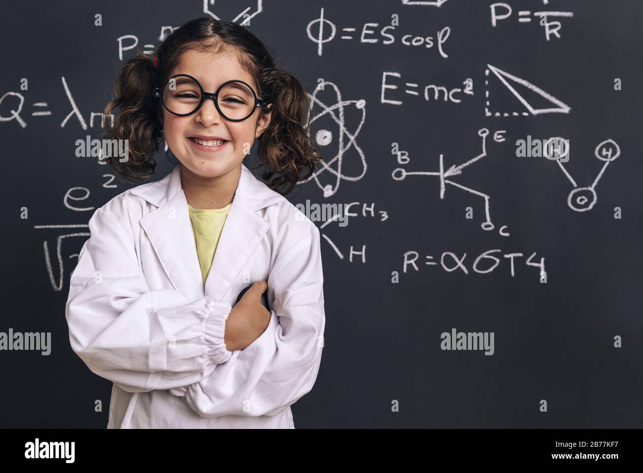 smiling little girl science student with glasses in lab coat on school blackboard background