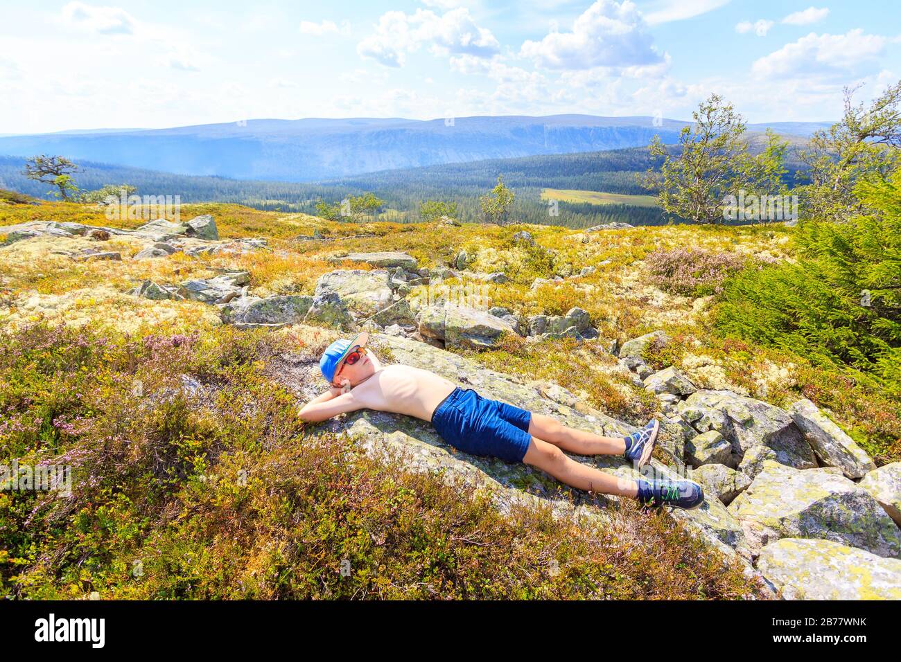 Kid resting lying down on the ground with t-shirt off, while hiking in Norway. Sunny weather, warm summer. Stock Photo