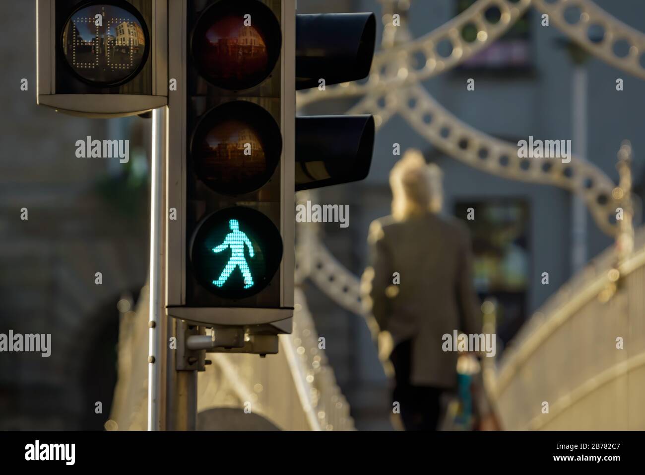 Selective focus on pedestrian traffic lights, buildings reflected in them, green walk sign. Blurred man walking on bridge in background Dublin Ireland Stock Photo