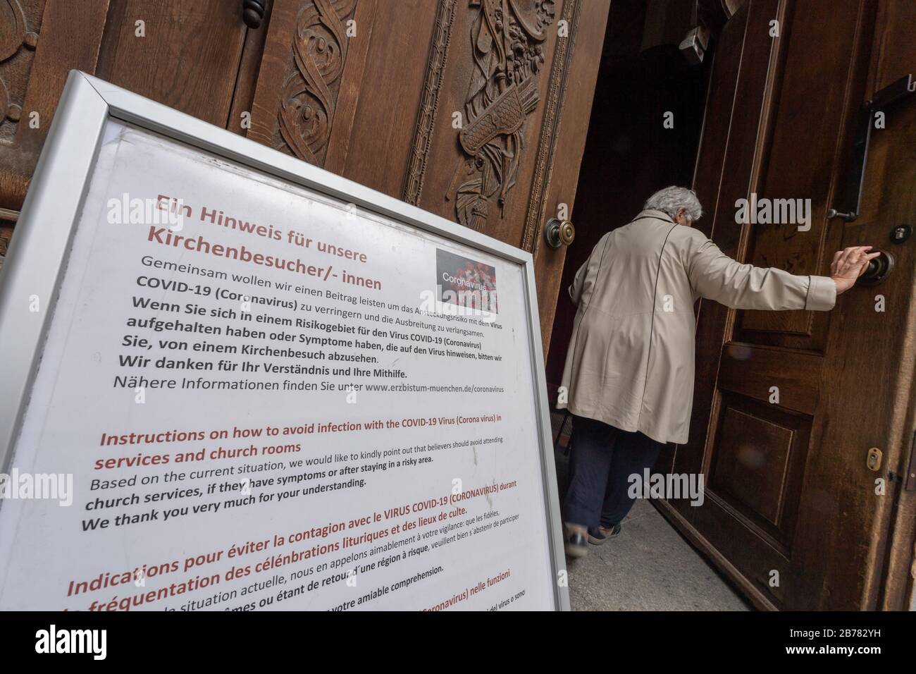Munich, Germany. 14th Mar, 2020. A woman walks through the entrance door of the Frauenkirche, in front of which an operator advises visitors not to go to church because of the coronavirus. Credit: Peter Kneffel/dpa/Alamy Live News Stock Photo