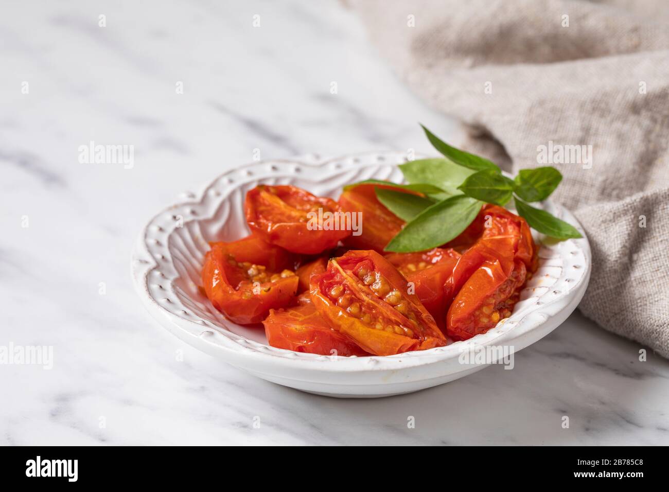 Sun dried tomatoes on a white plate, with a sprig of fresh basil as garment. The plate is on a white marble background, with a linen napkin on the sid Stock Photo