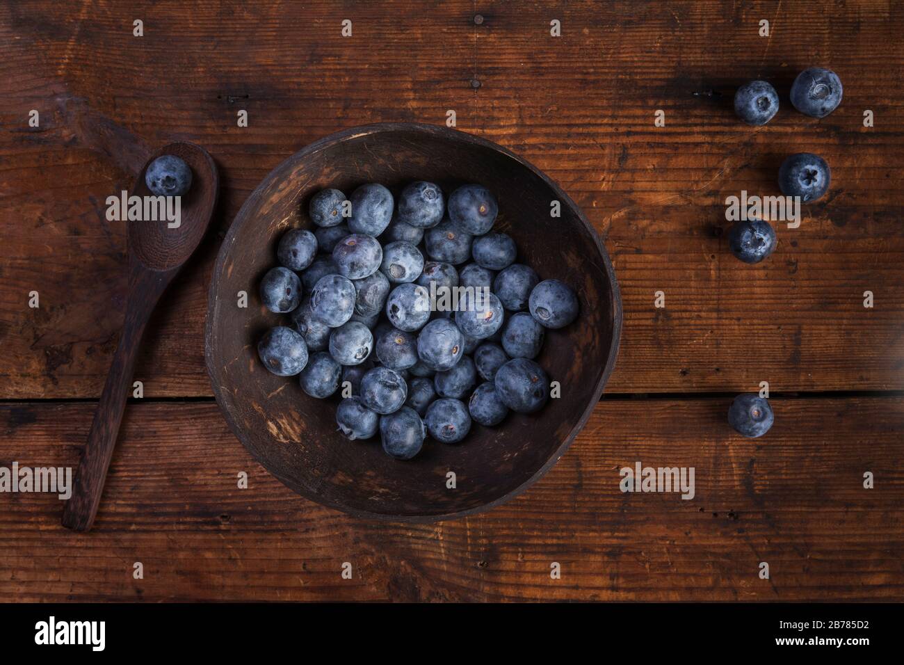 Blueberries or huckleberries in a coconut bowl with a wooden spoon. The bowl is on a rustic wooden table. Seen from above flat lay. There are some blu Stock Photo