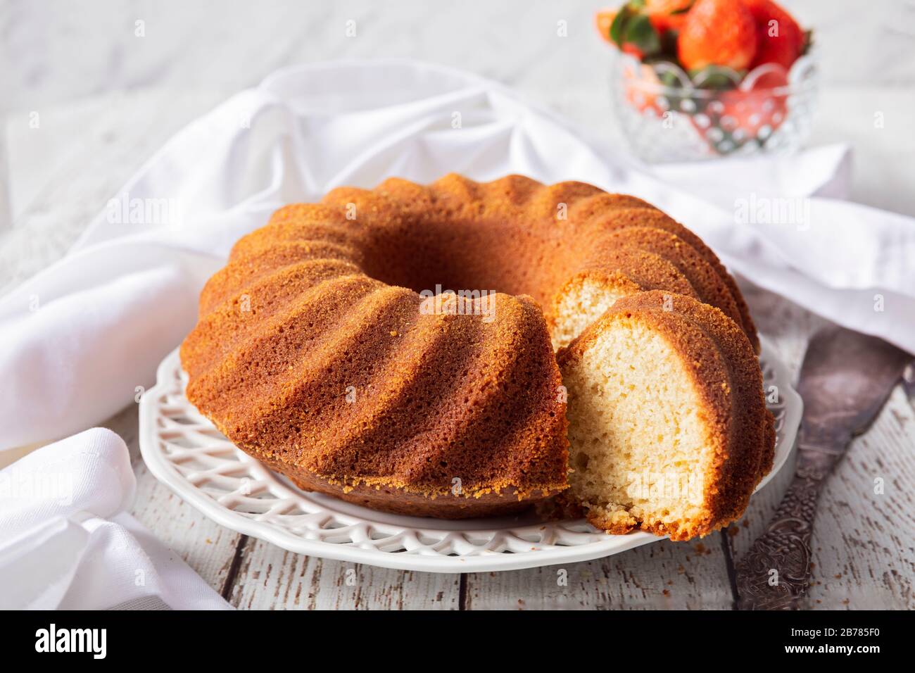 A white wooden table with a bundt cake , kugelhupf or sockerkaka. With a bowl of strawberries, a white napkin and a vintage cake cutter in the backgro Stock Photo