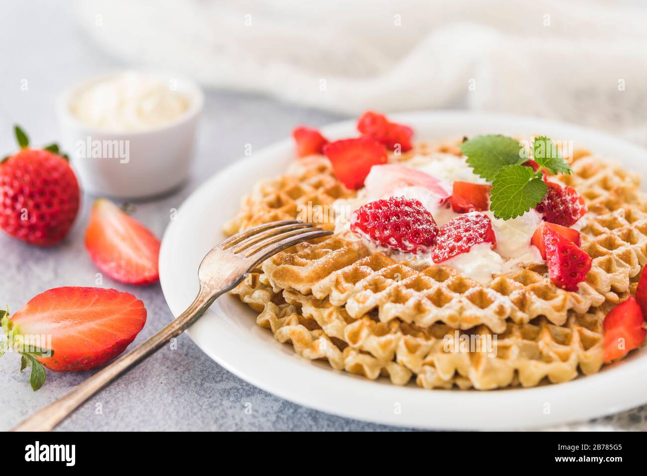 Swedish waffles with strawberries and whipped cream in a small bowl in the background. Next to the plate is a fork and whole and halved strawberries. Stock Photo