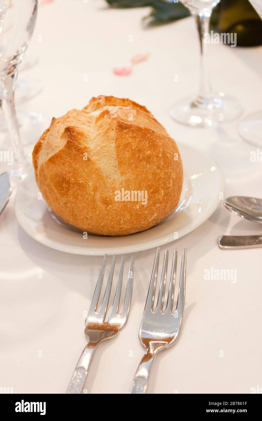 A bread bun on a plate on an elegant table with some cutlery and glass over a white cloth. Stock Photo