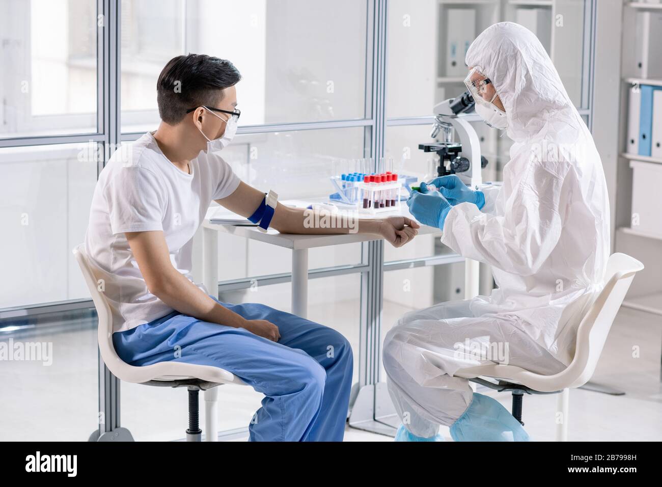 Worker of medical laboratory in protective workwear going to take blood of young Chinese man to diagnose disease Stock Photo