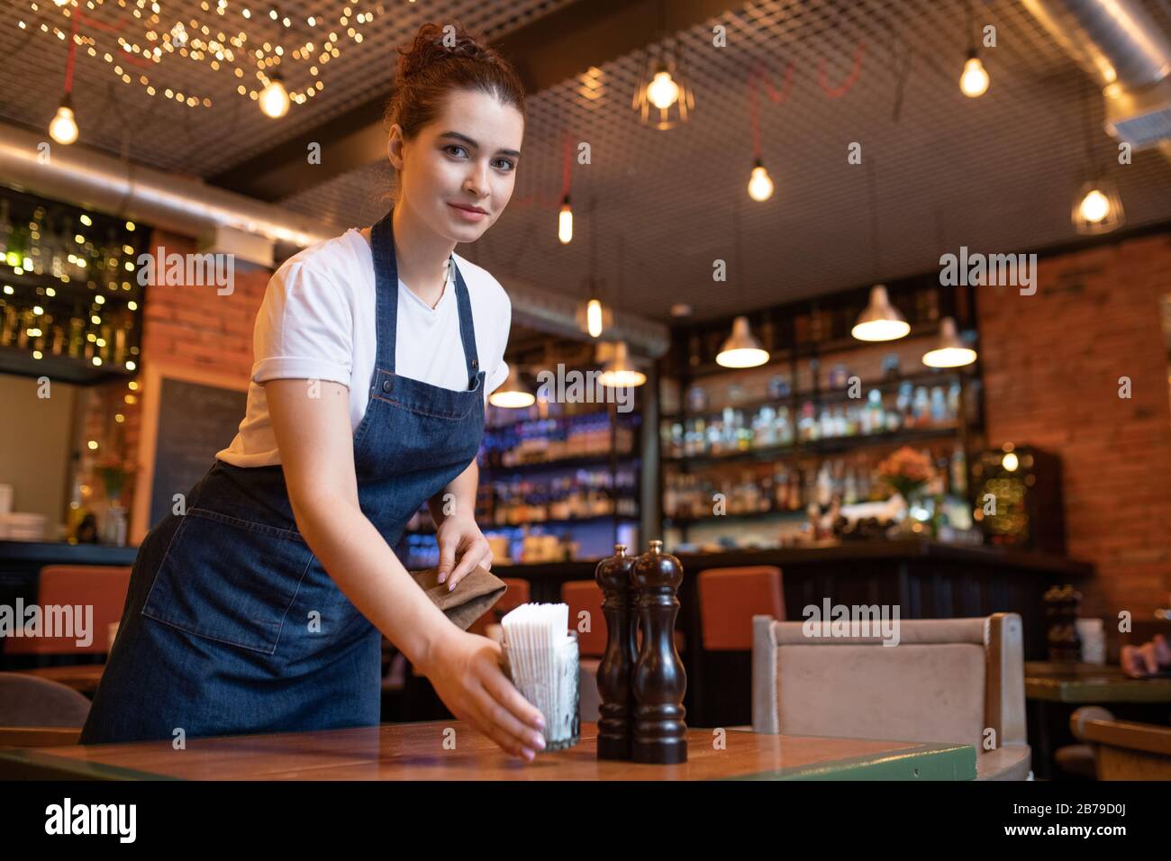 Pretty young waitress in workwear bending over table while putting glass with toothpicks and looking at you in cafe or restaurant Stock Photo