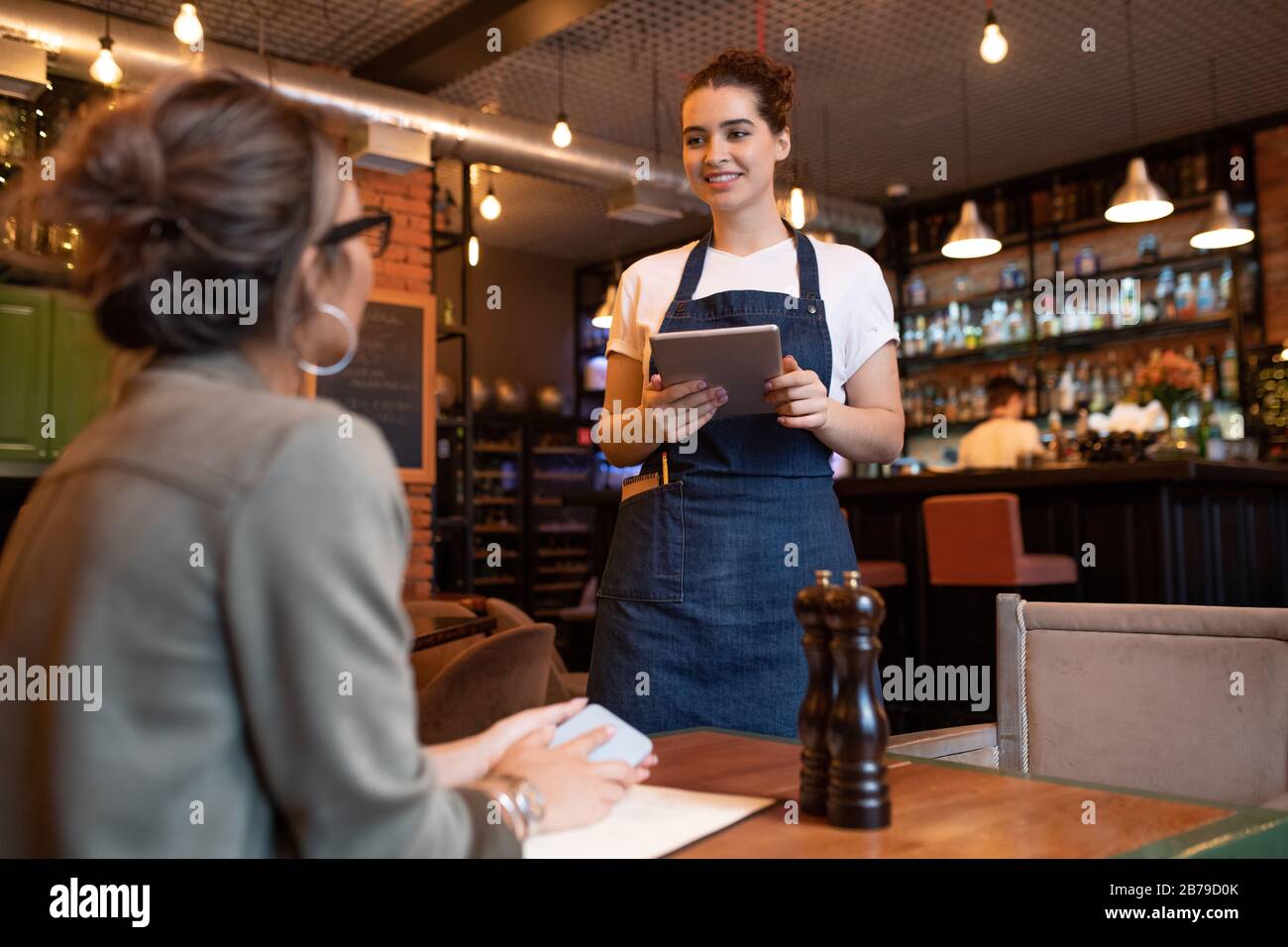Happy young waitress with digital tablet standing by one of tables in front of female guest and taking her order in cafe or restaurant Stock Photo
