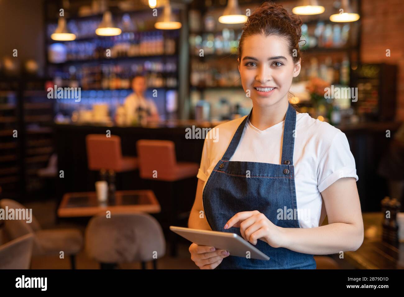 Young smiling waitress in apron and t-shirt standing in front of camera while using touchpad and meeting guests in cafe Stock Photo