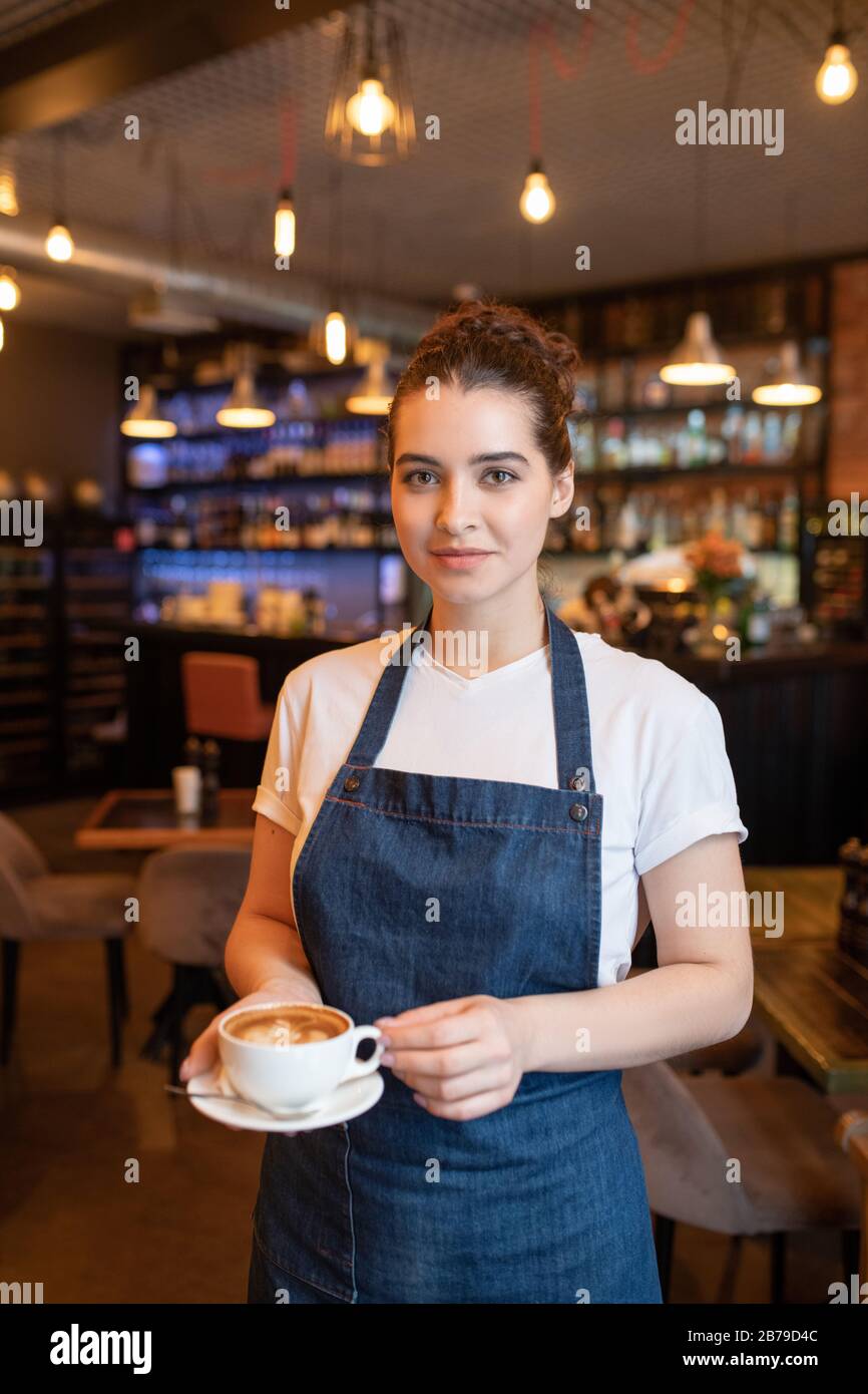 Pretty young brunette waitress with cup of cappuccino standing in front of camera on background of tables and chairs in cafe Stock Photo