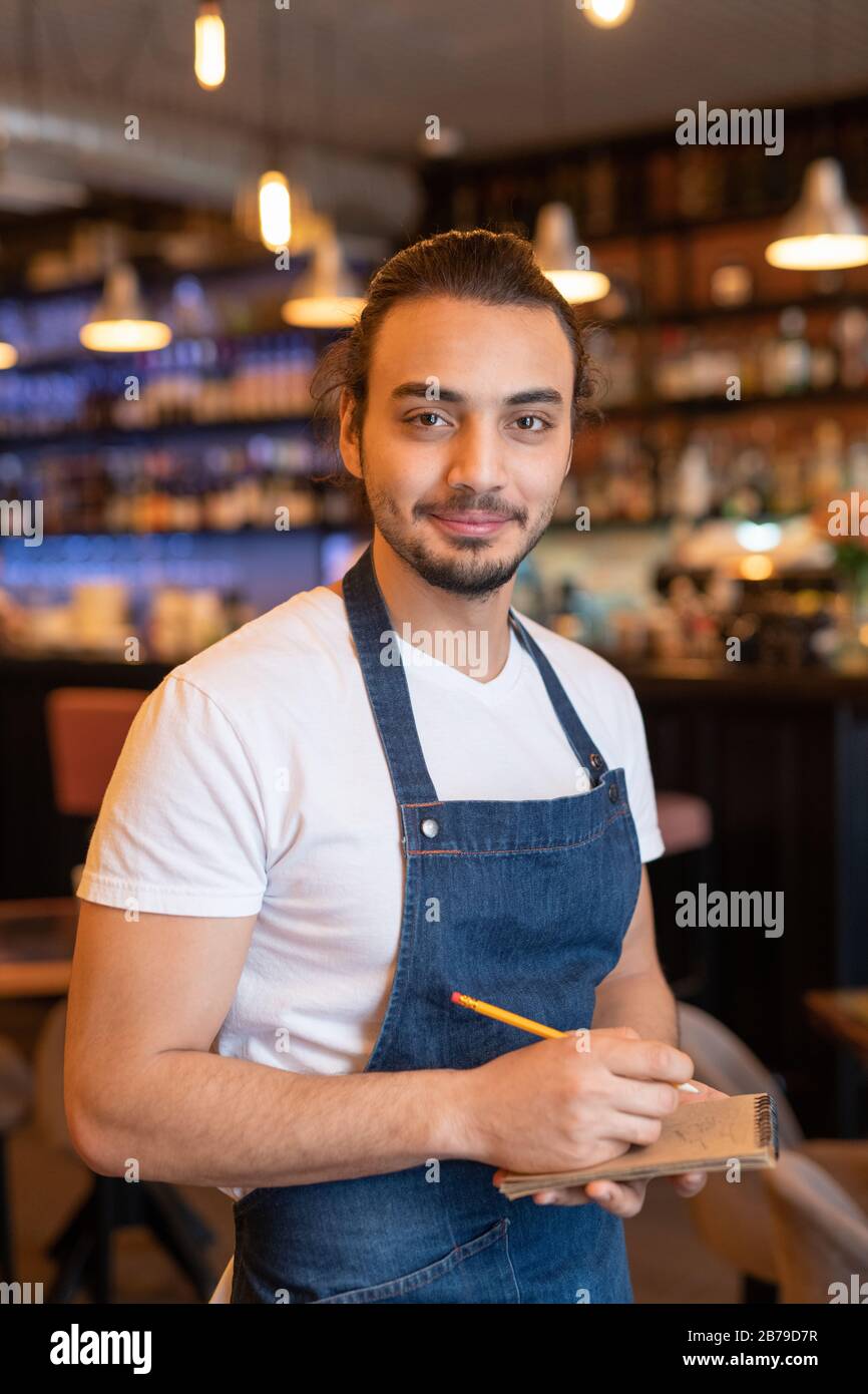 Young successful waiter in apron making notes in notepad while taking order of client during work in cafe or restaurant Stock Photo