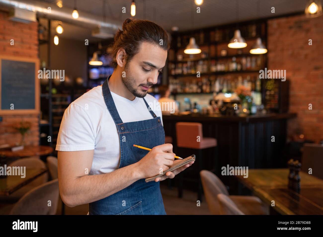 Young waiter in blue apron and white t-shirt writing down order of one of clients in notepad while standing in cafe or restaurant Stock Photo
