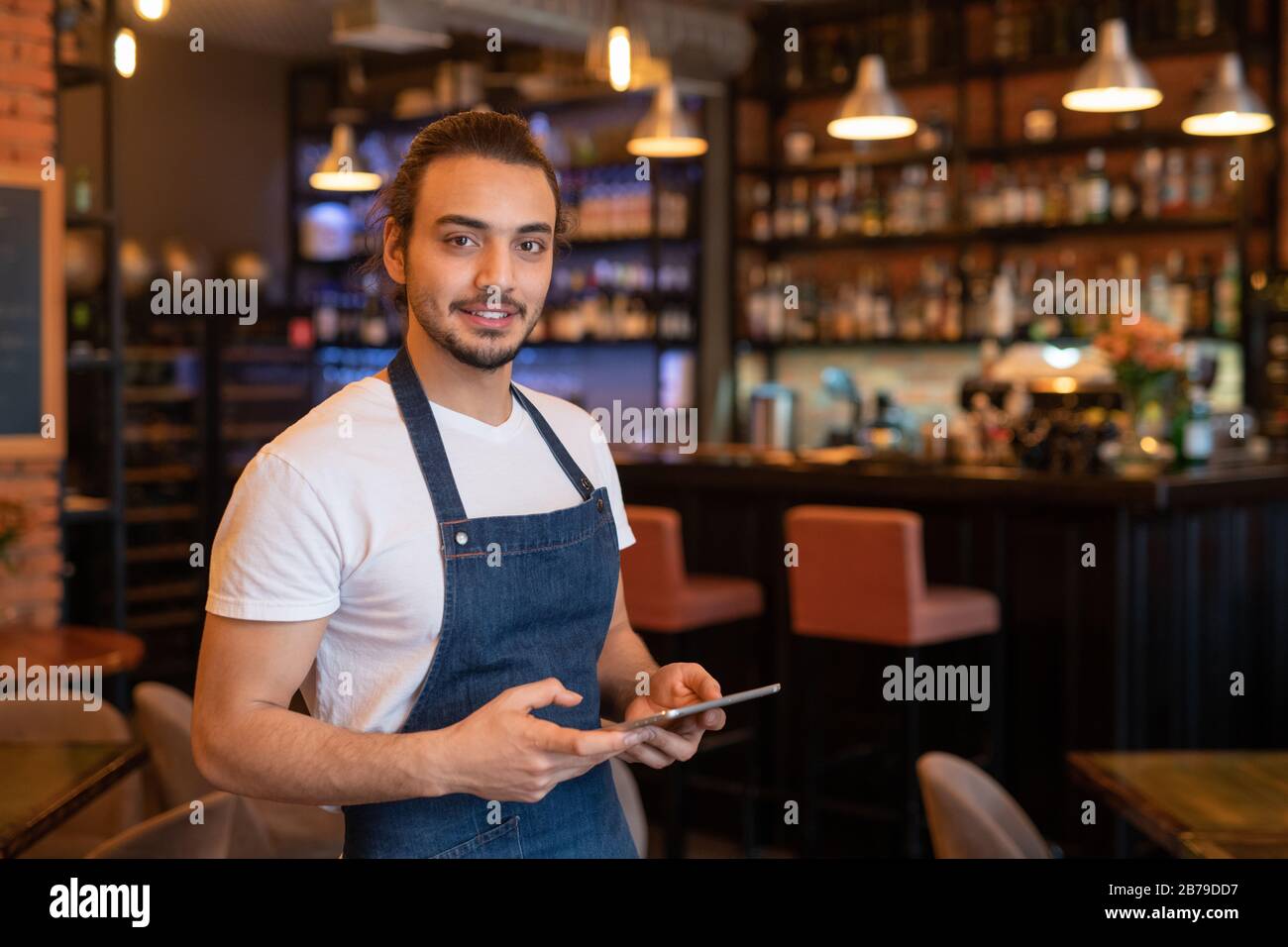 Contemporary young handsome waiter in apron using tablet while taking online orders of clients to come in the evening Stock Photo