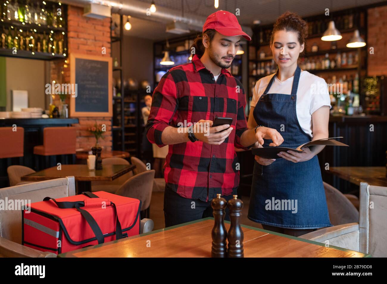 Young courier in flannel checking phone numbers and addresses of clients in special book held by waitress before deliver orders Stock Photo