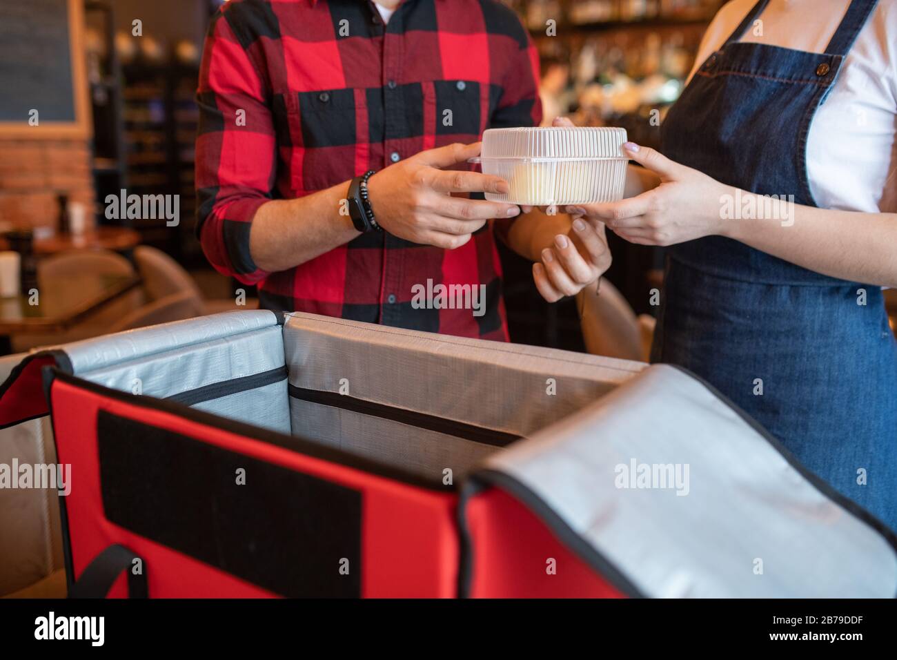 Waiter of cafe passing plastic container with order of client to courier over open big red bag while packing ordered food Stock Photo