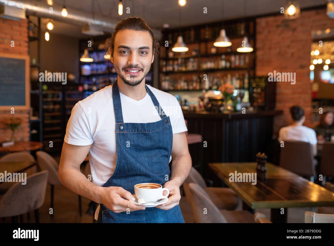 Young cheerful waiter in white t-shirt and blue apron holding cup of cappuccino on saucer while looking at you Stock Photo