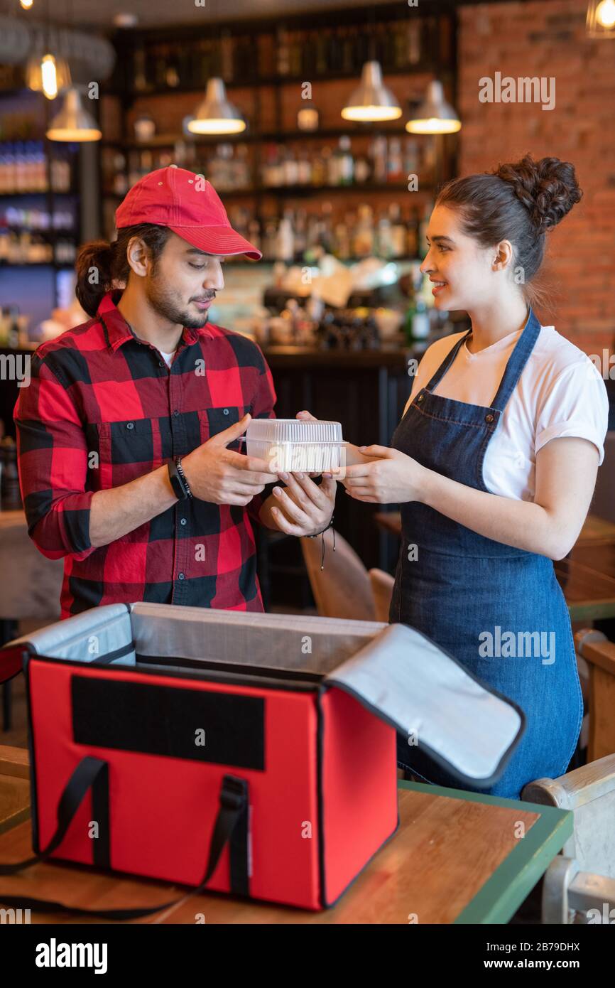 Young courier in workwear taking plastic container with food from hands of waitress helping him to pack orders of clients in cafe Stock Photo