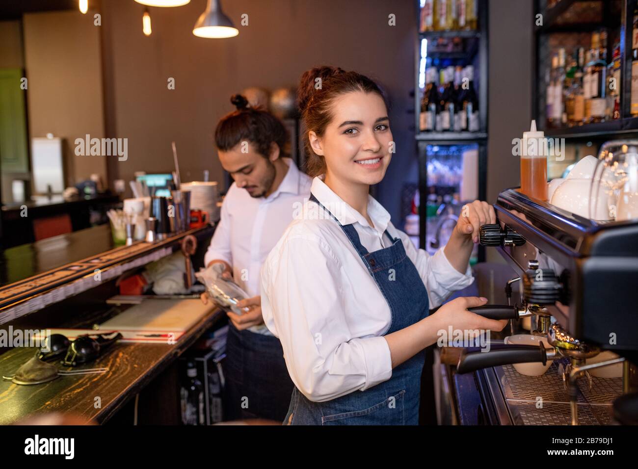 Happy young attractive waitress of cafe or restaurant preparing cappuccino by coffee machine with colleague on background Stock Photo