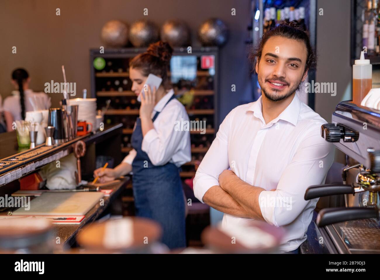 Happy young cross-armed waiter in white shirt standing in front of camera on background of colleague talking to client on the phone Stock Photo