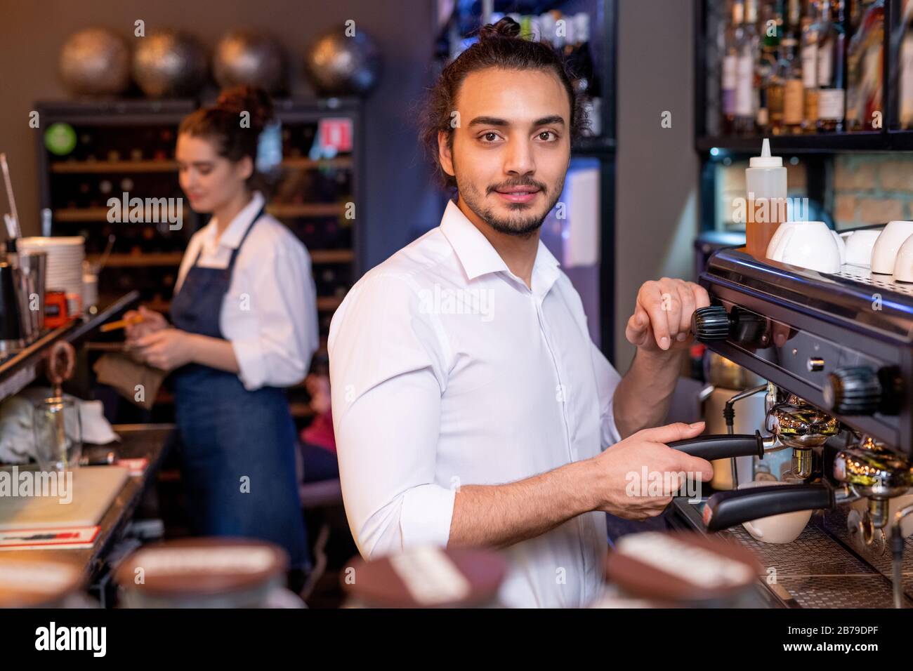 Young handsome waiter in white shirt preparing fresh cappuccino by coffee machine while his colleague wiping glass by workplace Stock Photo