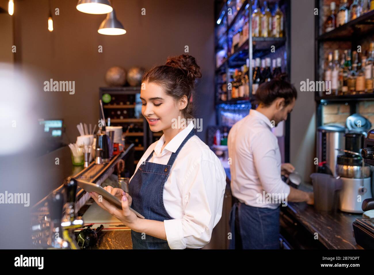 Pretty young waitress with tablet scrolling through online orders while her colleague preparing tea for guests on background Stock Photo