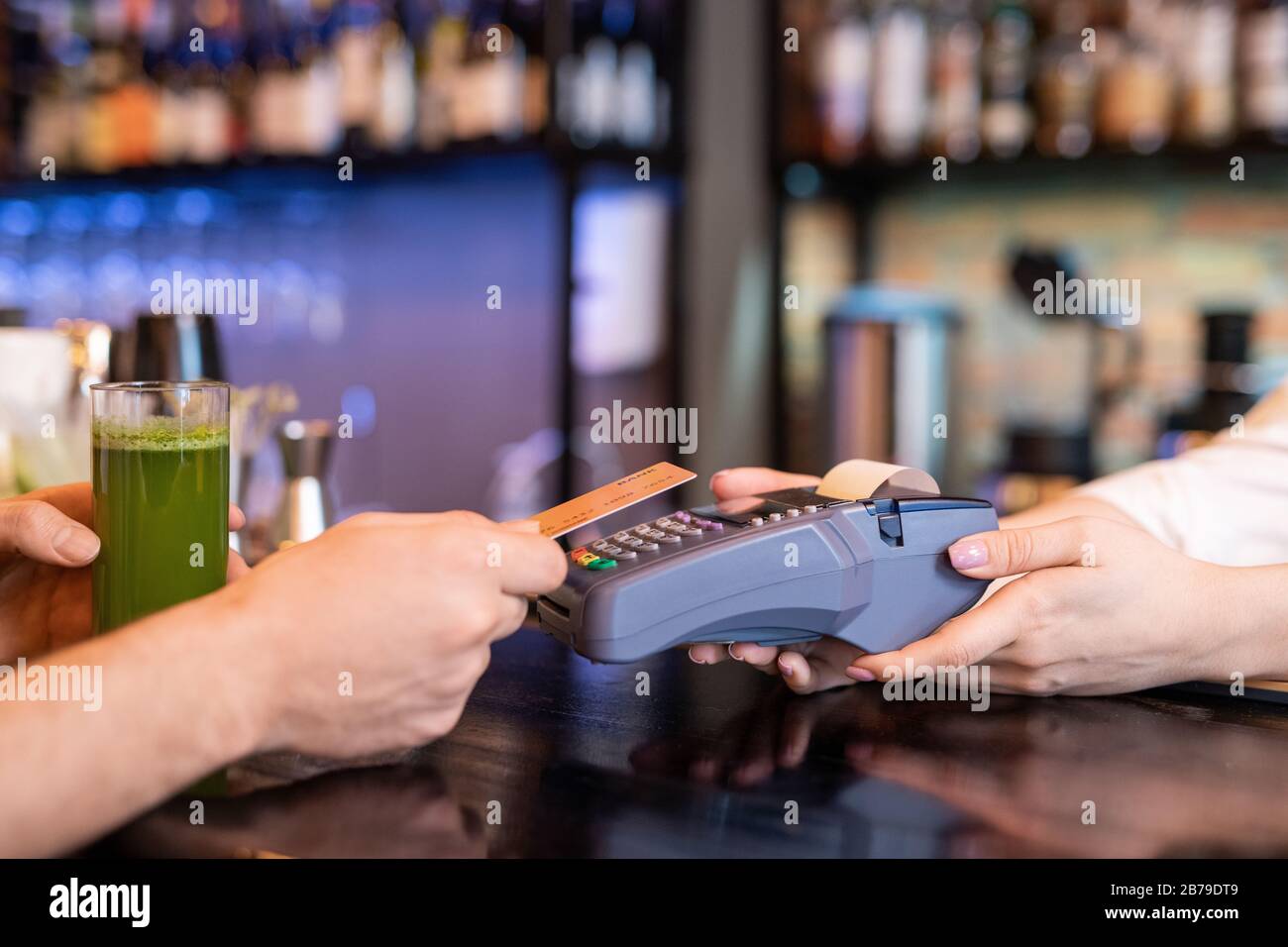 Hand of young man holding plastic card over payment machine held by waitress while paying for glass of fresh vegetable smoothie Stock Photo