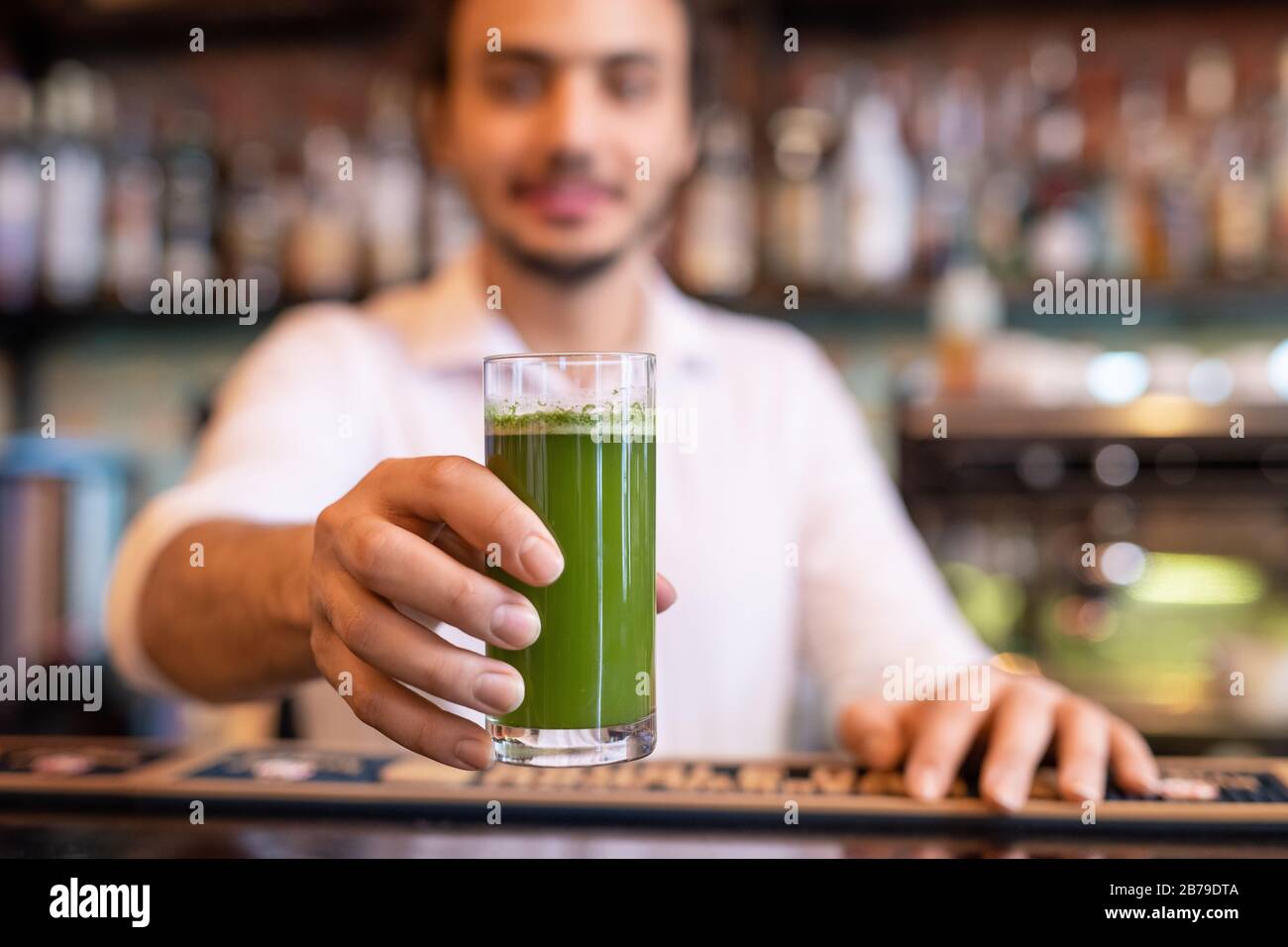 Hand of young barman holding glass of fresh vegetable smoothie over counter while giving it to one of guests of restaurant Stock Photo