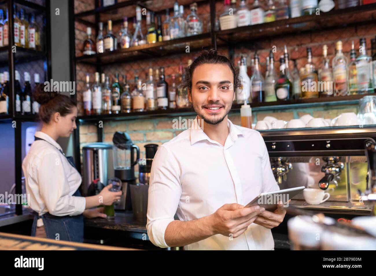 Young cheerful waiter with touchpad standing in front of camera while looking through online orders by workplace Stock Photo