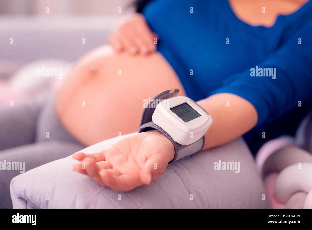 Pregnant woman monitoring her blood pressure at home with the medical device on her wrist and exposing her belly. Stock Photo