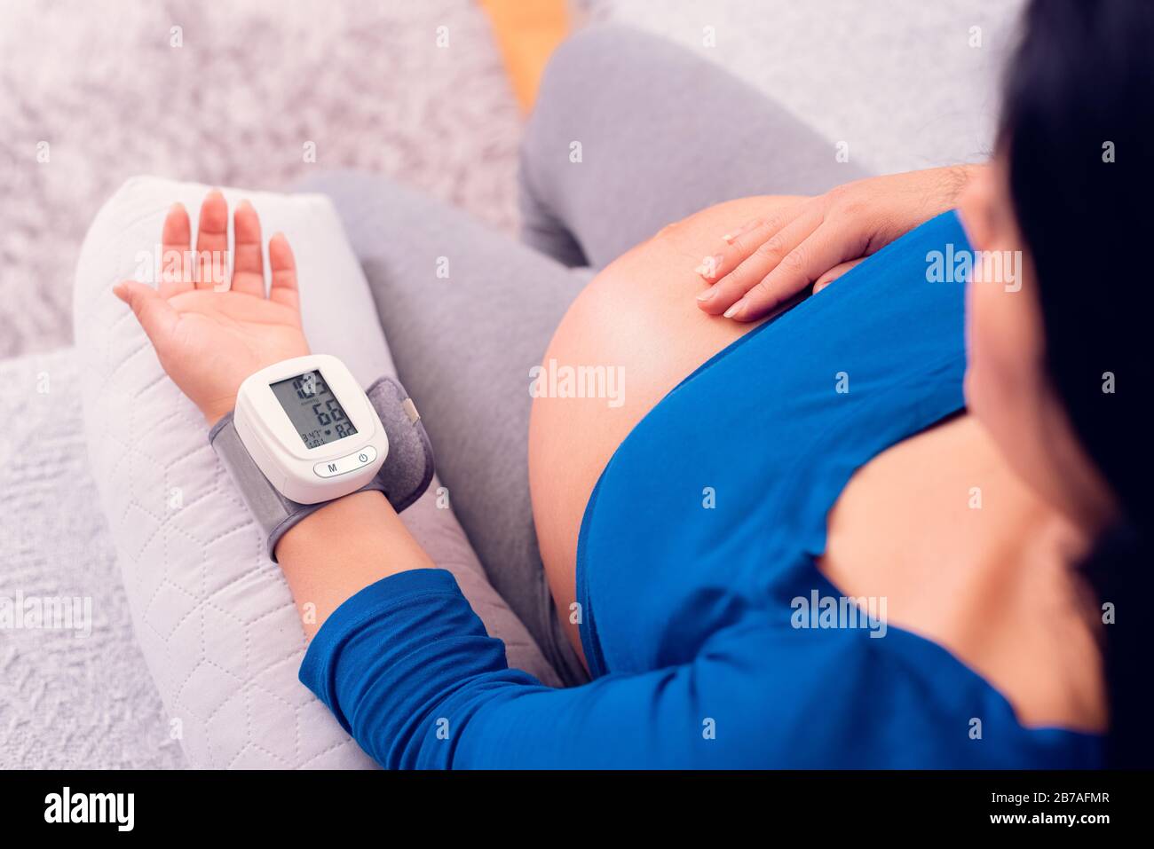 Pregnant woman monitoring her blood pressure at home with the medical device on her wrist and exposing her belly. Stock Photo