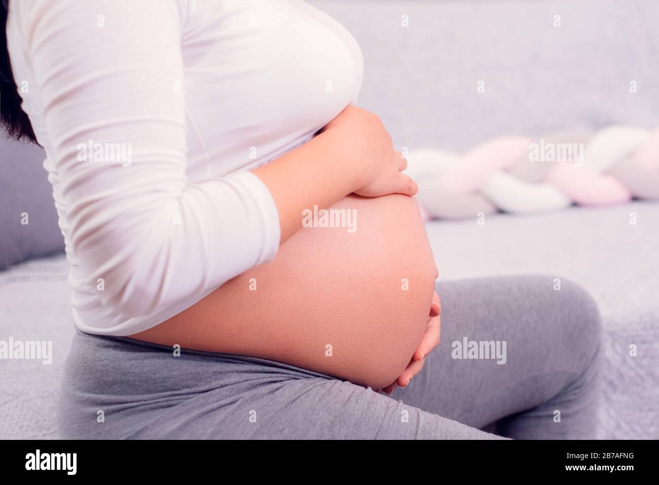 Pregnant woman sitting on the couch and holding her pregnant baby with two hands wearing withe top. Motherhood time. Stock Photo