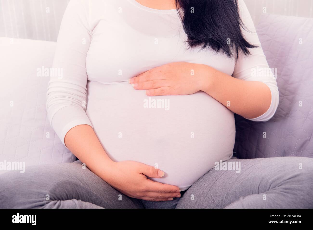 Pregnant woman sitting on the couch and holding her pregnant baby with two hands wearing withe top. Motherhood time. Stock Photo