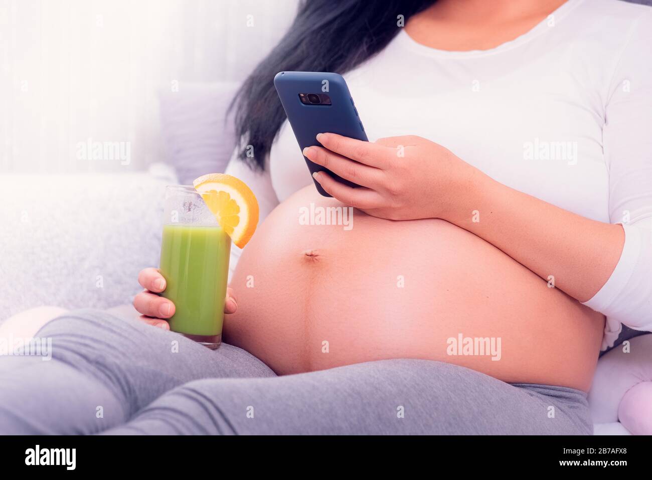 Pregnant woman using her smartphone while relaxing on the bed, drinking healthy green juice and exposing belly. Stock Photo
