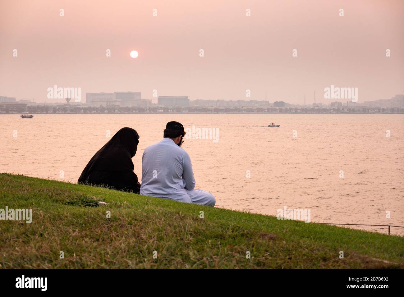 A Muslim man and woman in a burqa and thawb watch the sun set across the harbour in Doha, Qatar Stock Photo