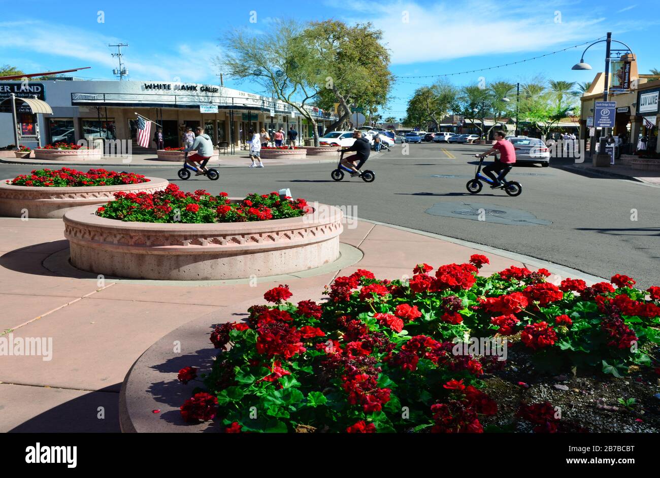 Three young men on electric scooters speed near red geraniums in foreground and galleries, restaurants and shopping district in Scottsdale, AZ Stock Photo